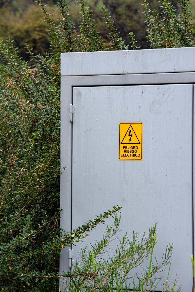 A gray electrical box surrounded by dense bushes with a warning sticker displayed prominently.